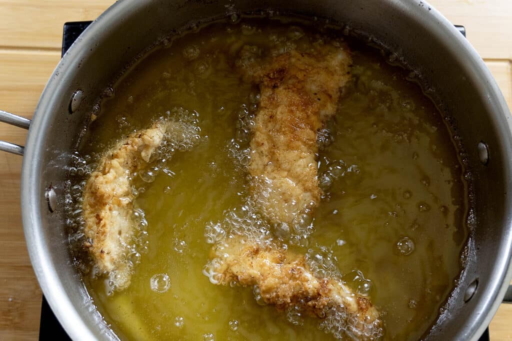 Three breaded chicken tenders are frying in hot oil inside a metal pot, with bubbles forming around the pieces as they cook. The pot is on a wooden surface.
