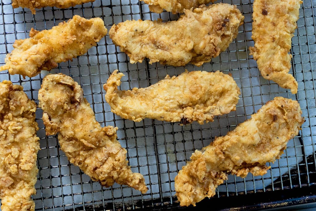 Several pieces of golden-brown fried chicken tenders rest on a metal cooling rack placed over a white surface.