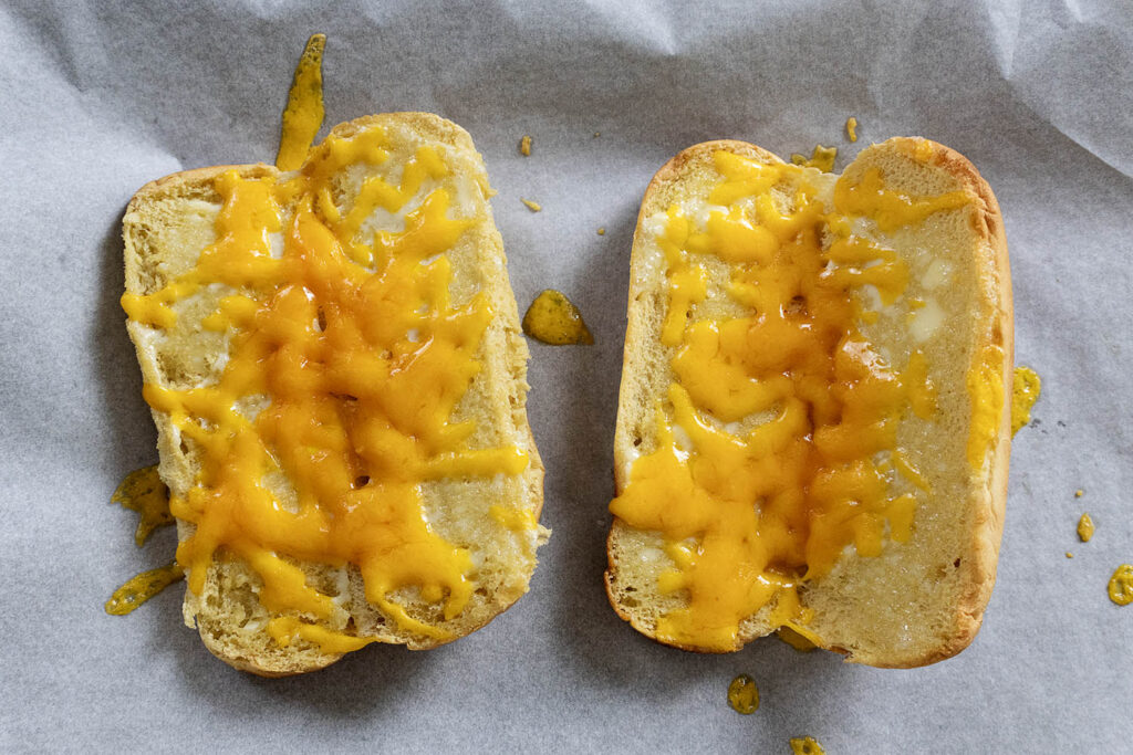 Two halves of a toasted bun topped with melted cheddar cheese are placed on a sheet of parchment paper. The cheese is slightly unevenly spread and melted, with some cheese drips on the paper.