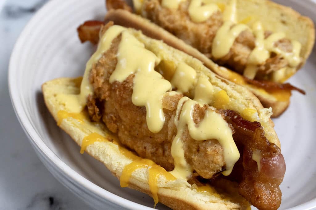 Two hot dog buns filled with crispy fried chicken strips, bacon, and melted cheese, topped with a generous drizzle of creamy yellow sauce, served on a white plate.