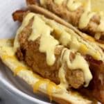 Two hot dog buns filled with crispy fried chicken strips, bacon, and melted cheese, topped with a generous drizzle of creamy yellow sauce, served on a white plate.