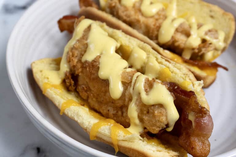 Two hot dog buns filled with crispy fried chicken strips, bacon, and melted cheese, topped with a generous drizzle of creamy yellow sauce, served on a white plate.