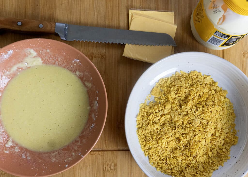 A bowl of batter, a bowl of crushed cornflakes, slices of processed cheese, a bread knife, and a jar of mayonnaise are arranged on a wooden surface.
