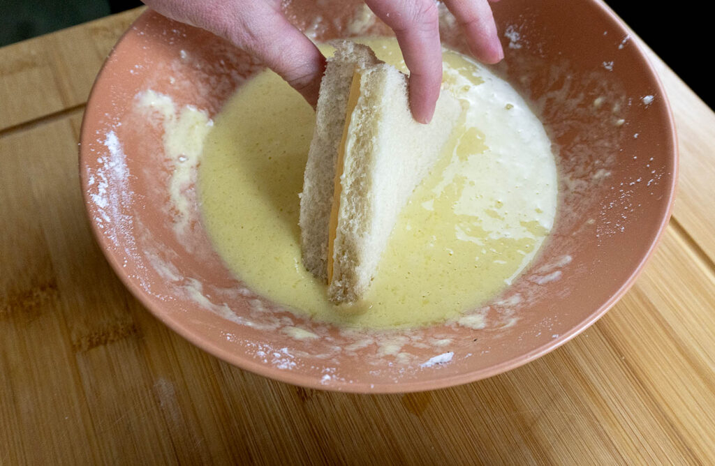 A hand dips two slices of white bread into a creamy yellow batter in an orange bowl on a wooden surface.