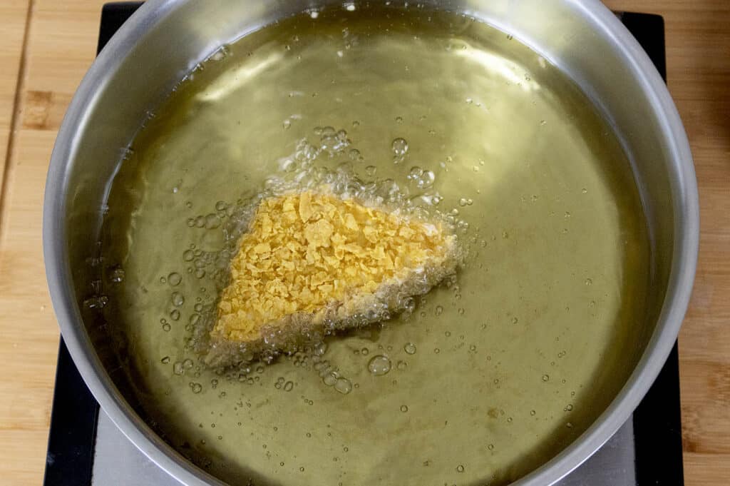 A triangular piece of food coated in crumbs is being deep-fried in hot oil inside a metal pan, with bubbles forming around it.