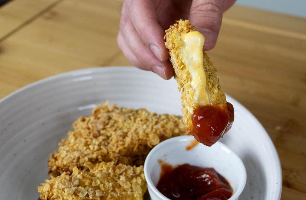 A hand holds a breaded, cheesy fried mozzarella stick, dipping it into a small bowl of red sauce, with more mozzarella sticks on a plate underneath.