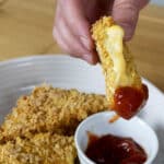 A hand holds a breaded, cheesy fried mozzarella stick, dipping it into a small bowl of red sauce, with more mozzarella sticks on a plate underneath.