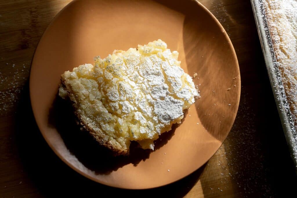 A square piece of lemon bar dusted with powdered sugar sits on an orange plate, with sunlight casting shadows across the plate and dessert.