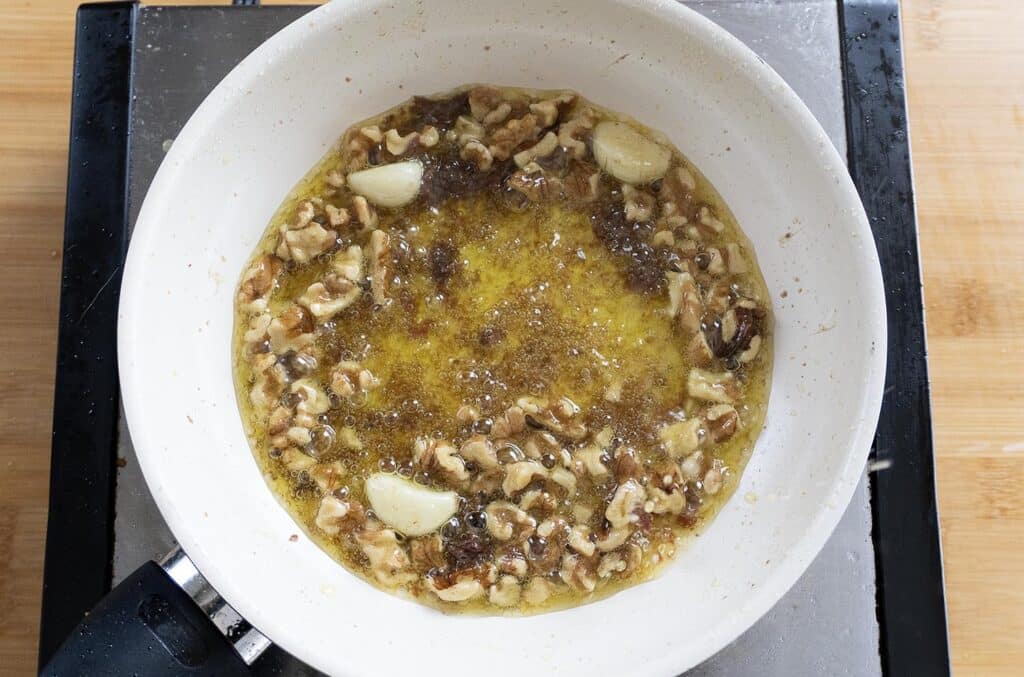 A white frying pan on a stovetop contains bubbling oil, walnut pieces, and several whole garlic cloves being sautéed. The pan sits on top of a black burner, and a wooden surface is visible underneath.