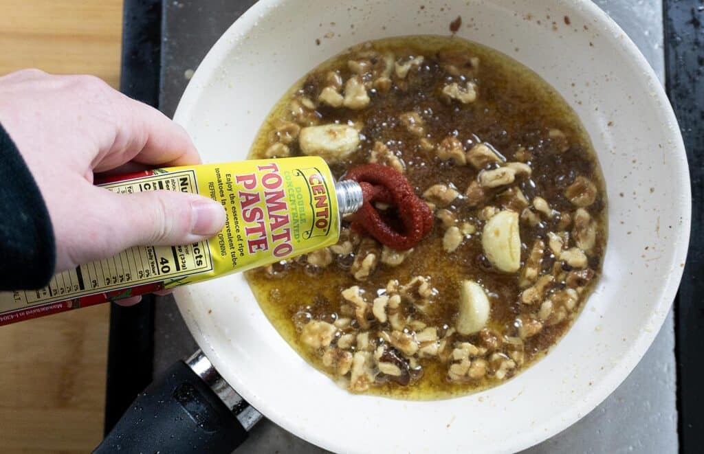 A hand squeezes tomato paste from a tube into a white pan with sizzling garlic, walnuts, and oil on a stovetop.