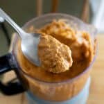 A close-up of a spoon holding a scoop of thick, orange-brown blended mixture above an open food processor filled with the same mixture, placed on a wooden surface.