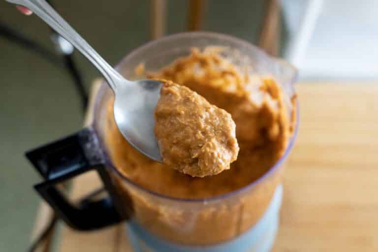 A close-up of a spoon holding a scoop of thick, orange-brown blended mixture above an open food processor filled with the same mixture, placed on a wooden surface.
