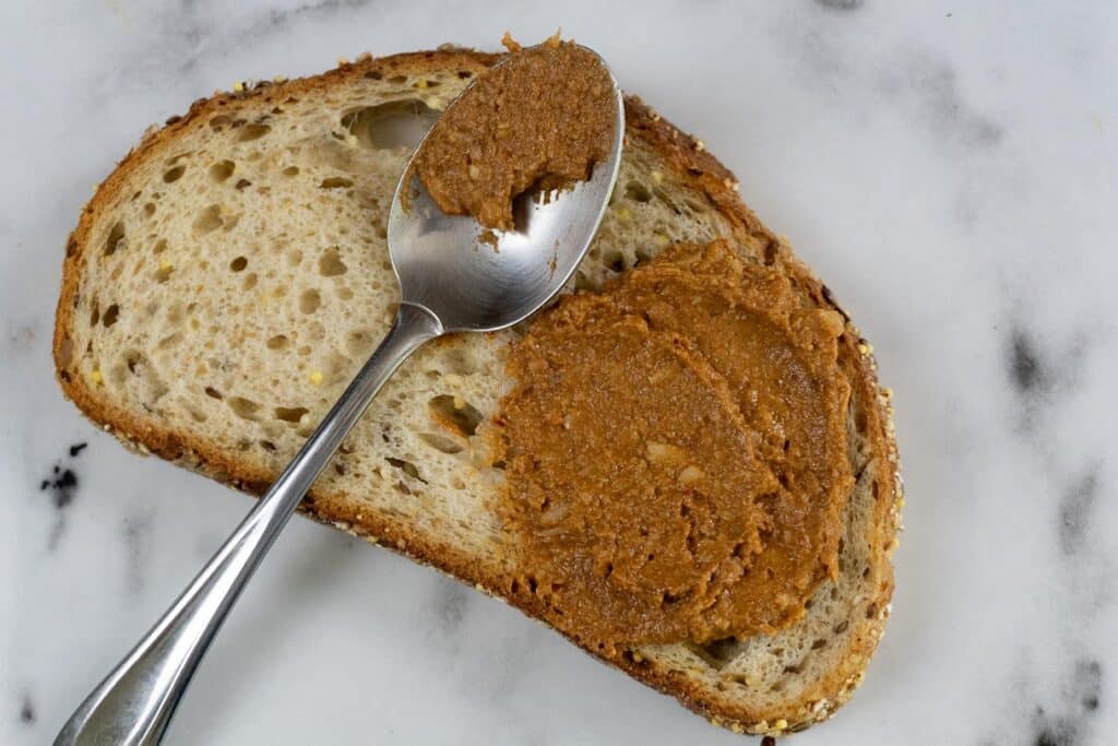 A slice of multigrain bread on a marble surface, topped with a spread of brown nut butter. A spoon with more nut butter rests on top of the bread.