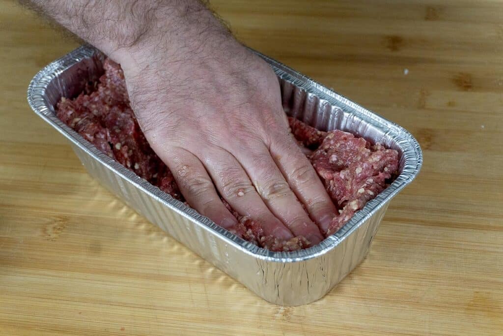 A hand presses raw ground meat mixture into a rectangular aluminum loaf pan on a wooden surface.