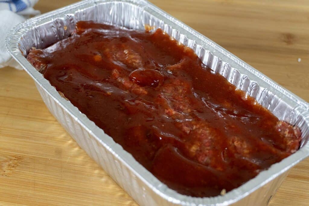 A rectangular aluminum tray filled with raw meatloaf mixture, topped with a layer of reddish-brown sauce, sitting on a wooden surface.