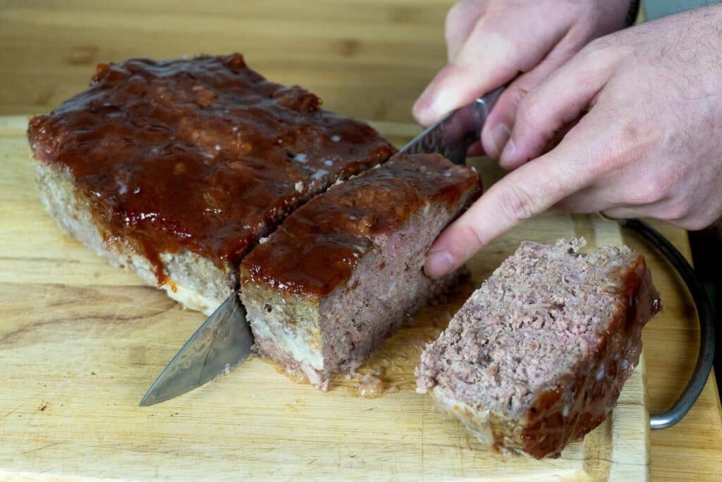 A person slices a glazed meatloaf on a wooden cutting board with a large knife, revealing the moist interior of the meatloaf.