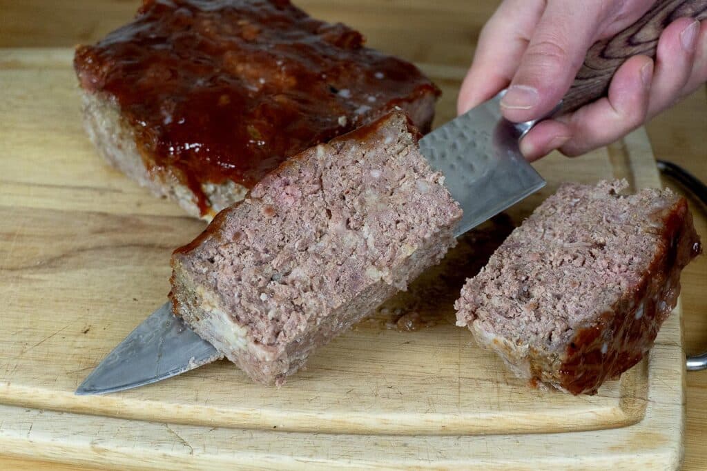 A hand slices a loaf of meatloaf with a large knife on a wooden cutting board. The meatloaf has a shiny glaze on top, and two thick slices are visible.