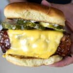 A close-up of a cheeseburger with a thick grilled patty, melted cheese, pickles, onions, and a soft hamburger bun, being held in someone’s hands.