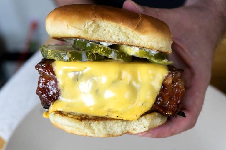 A close-up of a smoked meatloaf sandwich held in two hands, featuring melted cheddar cheese, grilled meat, pickles, chopped onions, and a soft, golden bun.