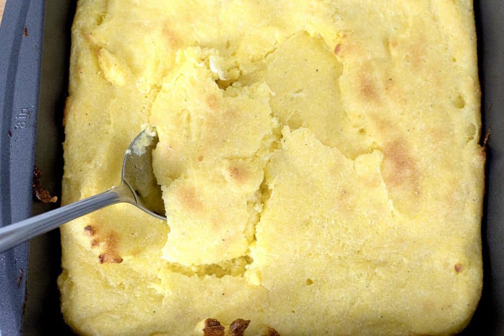 A metal spoon scoops a portion of a golden, baked cornbread from a rectangular baking pan. The cornbread has a slightly browned top and a soft, crumbly texture.