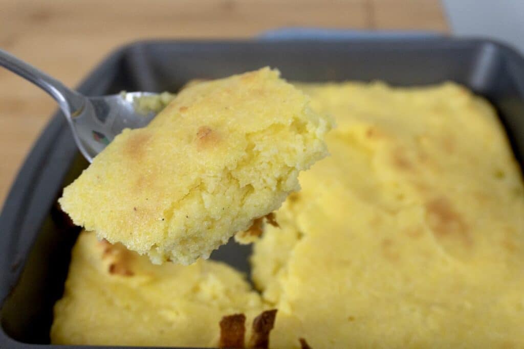 A close-up of a fork holding a piece of golden, fluffy cornbread above a baking pan filled with the remaining cornbread. The cornbread has a slightly browned top and a moist, crumbly texture.