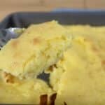 A close-up of a fork holding a piece of golden, fluffy cornbread above a baking pan filled with the remaining cornbread. The cornbread has a slightly browned top and a moist, crumbly texture.