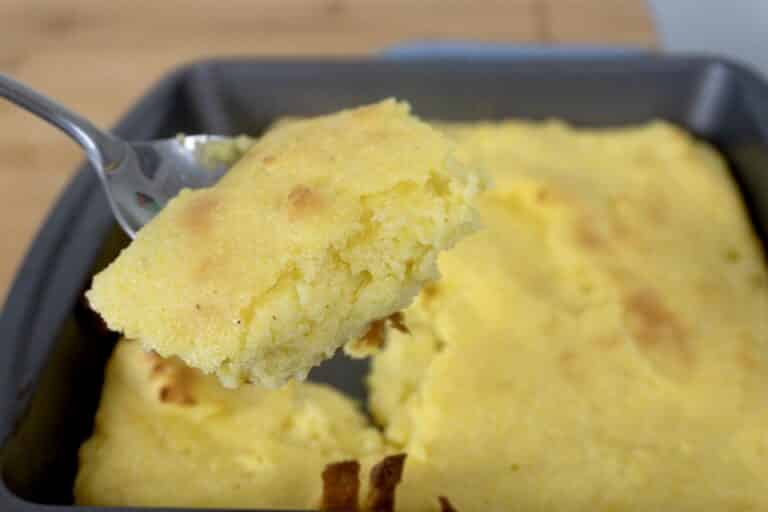 A close-up of a fork holding a piece of golden, fluffy cornbread above a baking pan filled with the remaining cornbread. The cornbread has a slightly browned top and a moist, crumbly texture.