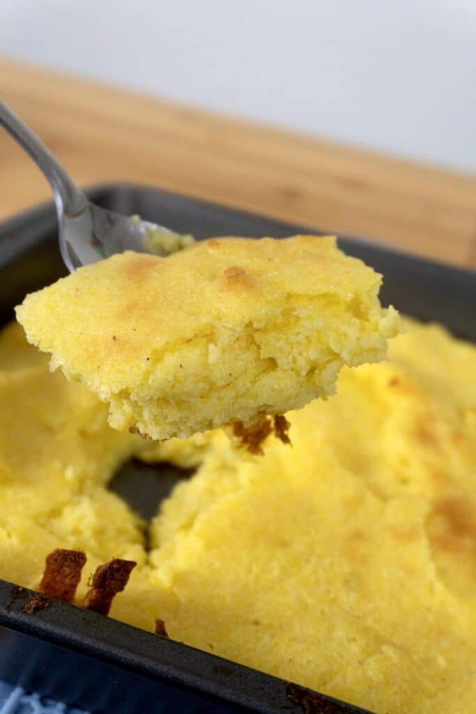 A close-up of a spoon holding a portion of golden, fluffy cornbread above a baking pan filled with more cornbread.