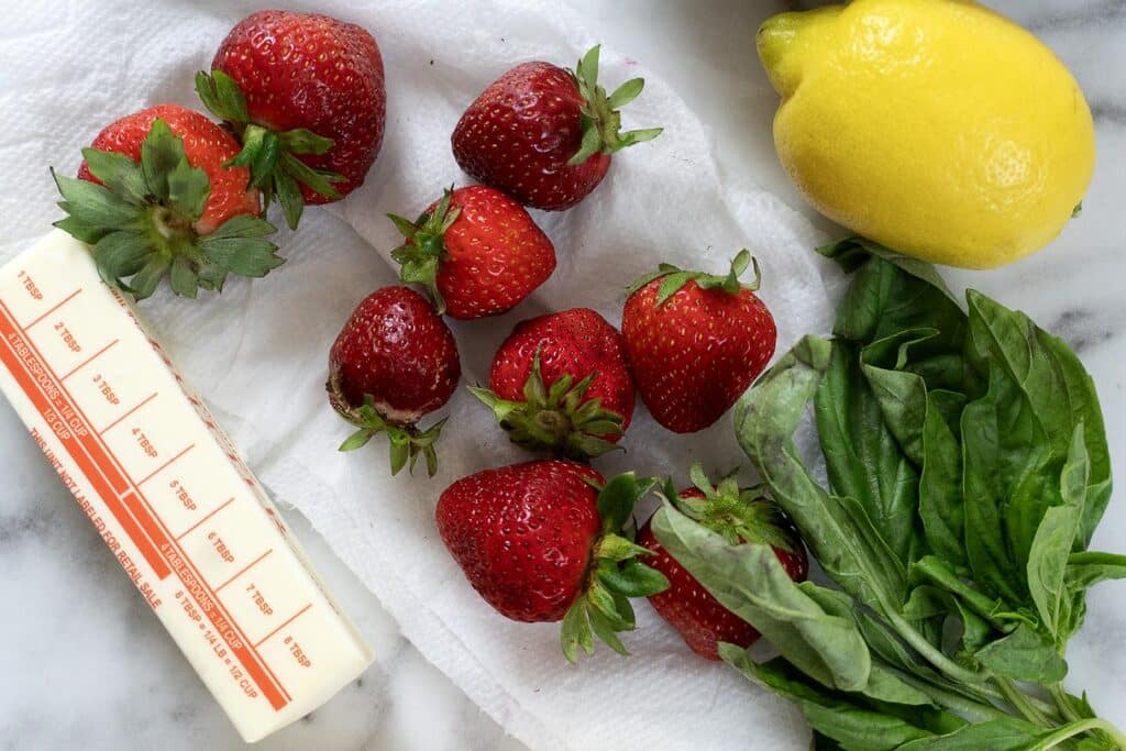 Fresh strawberries, a stick of butter, a bunch of basil leaves, and a whole lemon are arranged on a paper towel atop a marble surface.