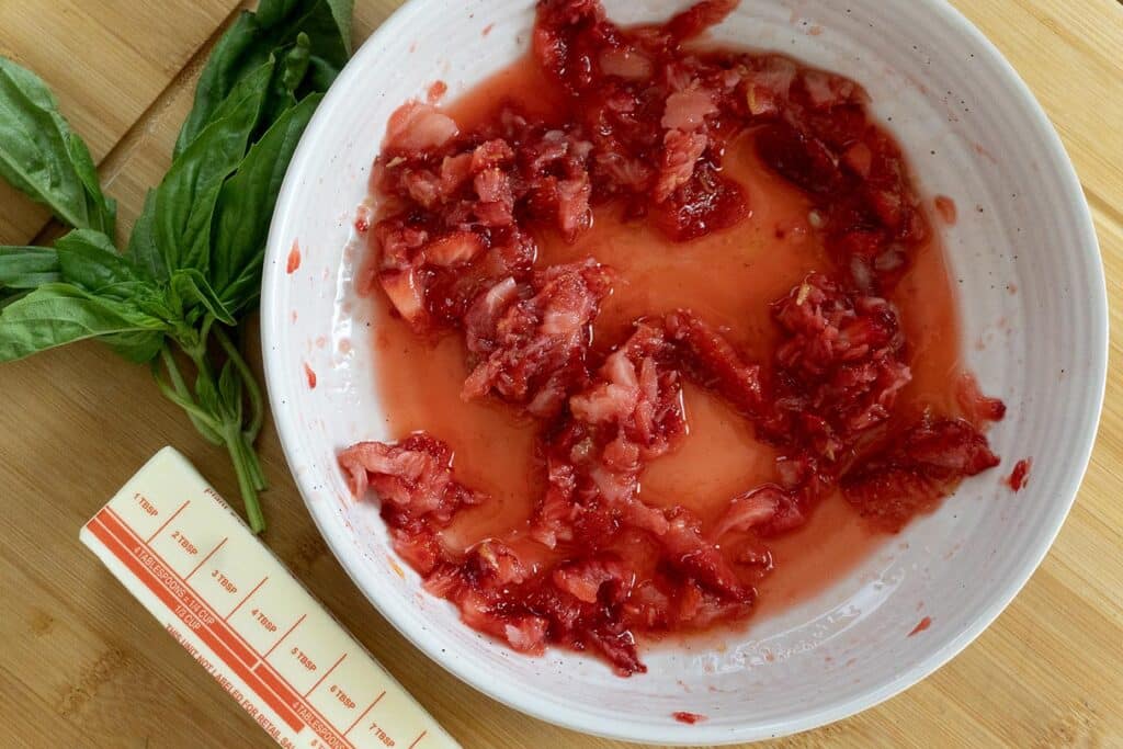 A white bowl with mashed strawberries and juice, next to a stick of butter with measurement markings and a small bunch of fresh basil leaves, all on a wooden surface.