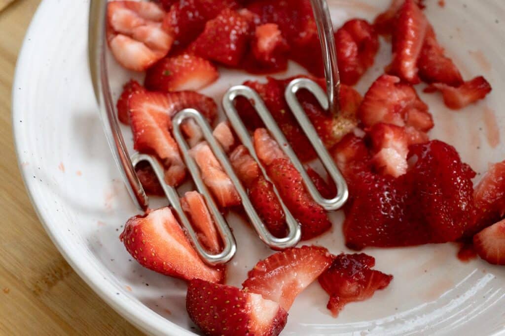 A metal potato masher presses down on sliced strawberries in a white bowl, partially crushing the fruit.