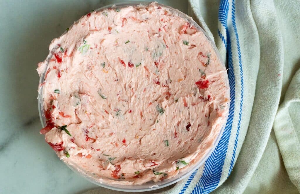 A bowl of pink, creamy spread with visible bits of herbs and red peppers sits on a marble surface next to a beige cloth with blue stripes.