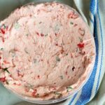A bowl of pink, creamy spread with visible bits of herbs and red peppers sits on a marble surface next to a beige cloth with blue stripes.