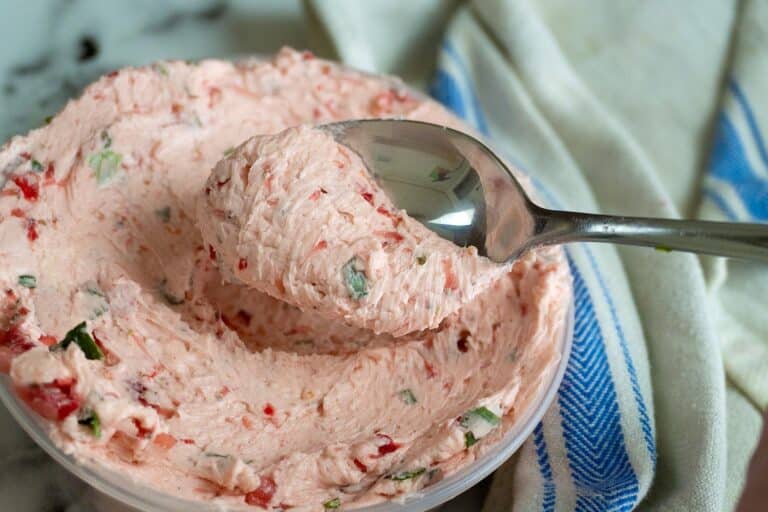 A metal spoon scoops up a creamy, pink spread with visible green and red bits from a bowl. A white and blue striped cloth is next to the bowl on a marble surface.