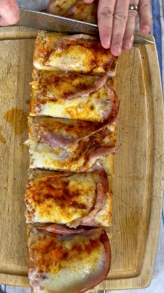 A hand holds a knife, cutting a rectangular baked dish with melted cheese and ham into six slices on a wooden cutting board.