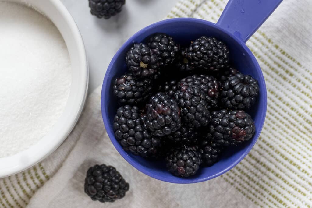 A blue measuring cup filled with blackberries sits on a striped cloth next to a white bowl of granulated sugar and a few loose blackberries.