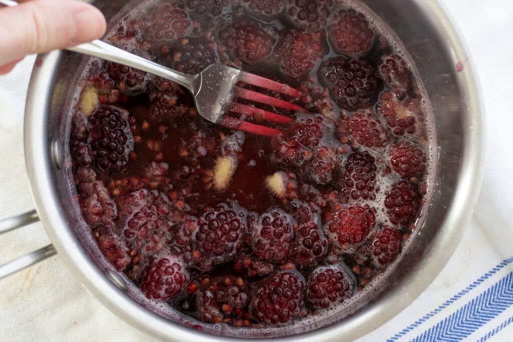 A metal saucepan filled with simmering blackberries, sugar, and liquid, being stirred with a fork; the mixture appears to be cooking on a white and blue towel.