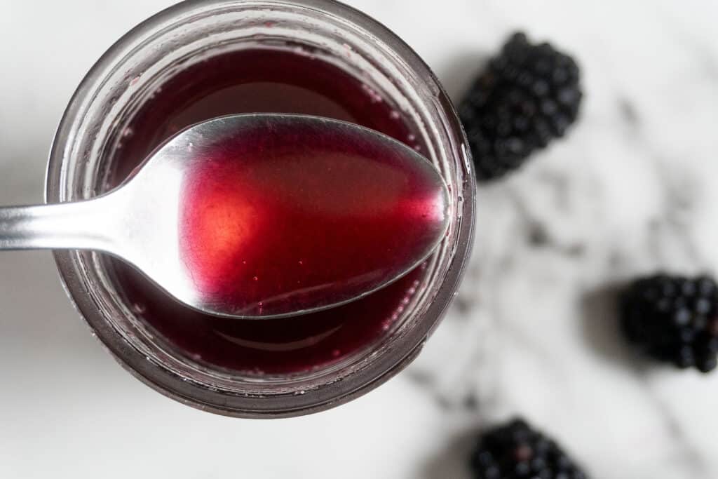 A close-up of a spoon holding red berry syrup above a jar, with two blackberries blurred in the background on a white surface.