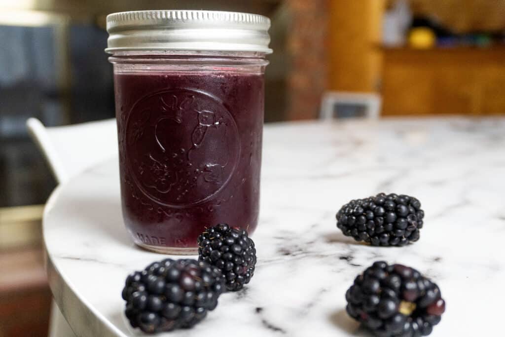 A glass jar filled with dark purple blackberry jam sits on a marble table, with four fresh blackberries arranged around it.