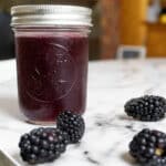 A glass jar filled with dark purple blackberry jam sits on a marble table, with four fresh blackberries arranged around it.
