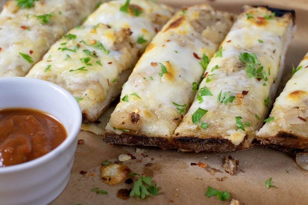 Sliced cheesy garlic bread topped with chopped parsley and red pepper flakes, served on parchment paper with a small white cup of marinara sauce—an irresistible cheesy bread inspired by our favorite garlic bread recipe.
