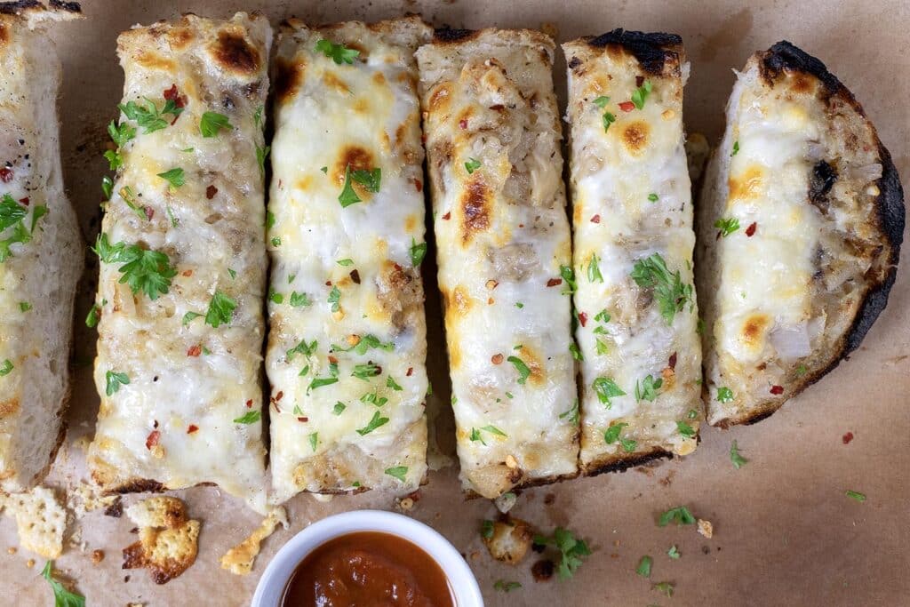 Sliced cheesy garlic bread topped with melted cheese, herbs, and red pepper flakes, served on brown parchment paper with a small cup of marinara dipping sauce.
