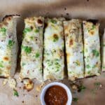 Sliced cheesy garlic bread topped with herbs on parchment paper, served with a small bowl of red dipping sauce.