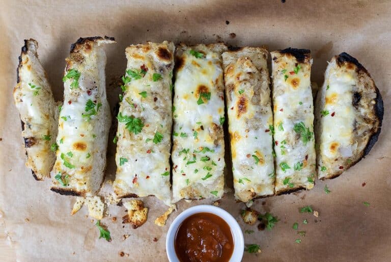 Sliced cheesy garlic bread topped with herbs on parchment paper, served with a small bowl of red dipping sauce.