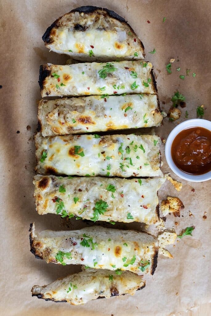 A sliced cheesy garlic bread, made from a classic garlic bread recipe and topped with herbs, is laid on parchment paper, with a small cup of marinara sauce on the side.
