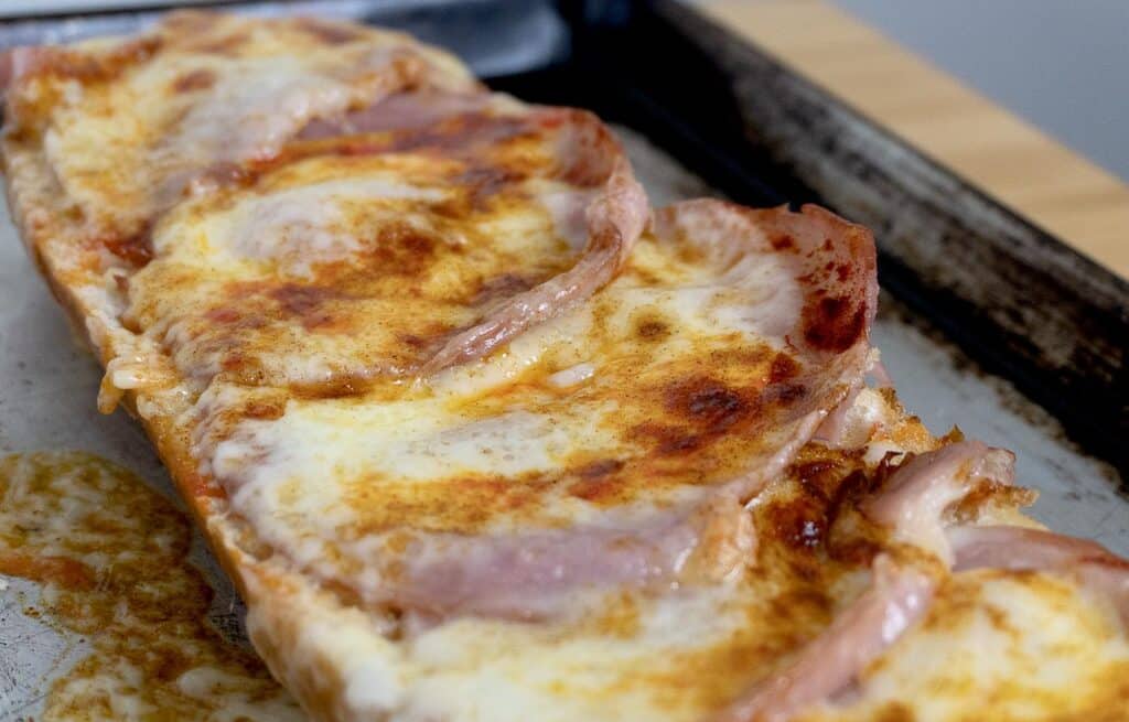 A close-up of a slice of a Gerber Sandwich topped with melted cheese and several strips of ham, resting on a metal baking sheet.