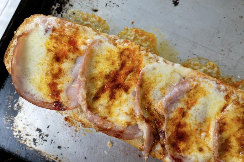 A toasted open-faced Gerber sandwich topped with melted cheese, ham slices, and a sprinkle of paprika sits on a metal baking sheet with spots of melted cheese around it.