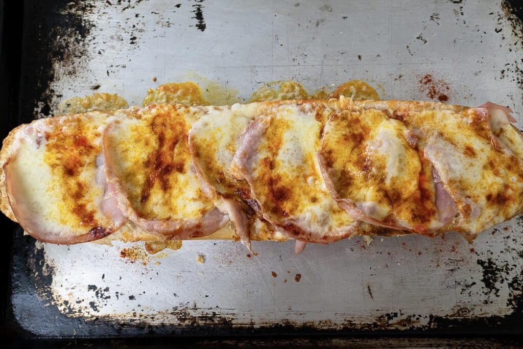 A toasted baguette topped with melted cheese, slices of ham, and sprinkled paprika on a worn metal baking sheet—just like a classic Gerber sandwich recipe.