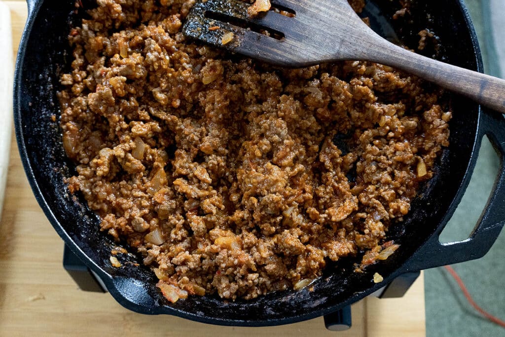 Seasoned ground beef with diced onions cooking in a black cast iron skillet, being stirred with a wooden spatula on a wooden surface.