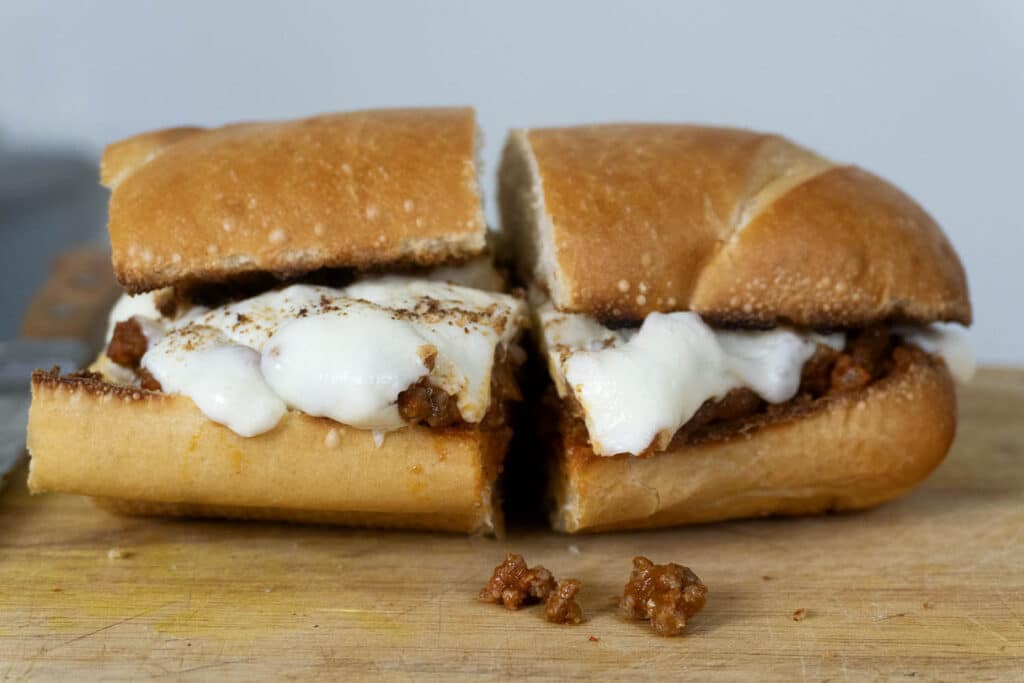 A hoagie roll sandwich filled with ground meat and topped with melted white cheese, sliced in half and placed on a wooden surface with a few crumbs nearby.